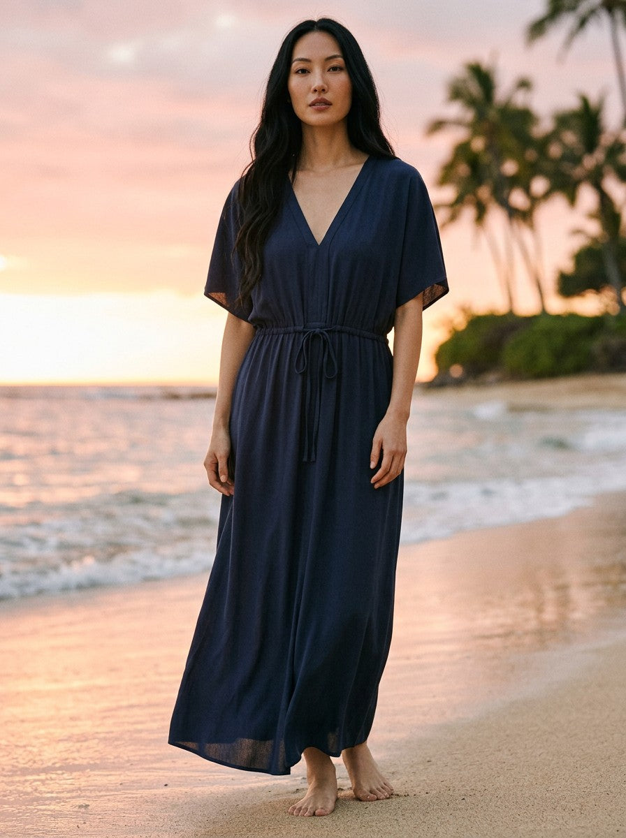 Antique Rose Boutique navy blue ECOVERO kaftan dress with V-neck and tie waist worn by a woman on a beach at sunset.