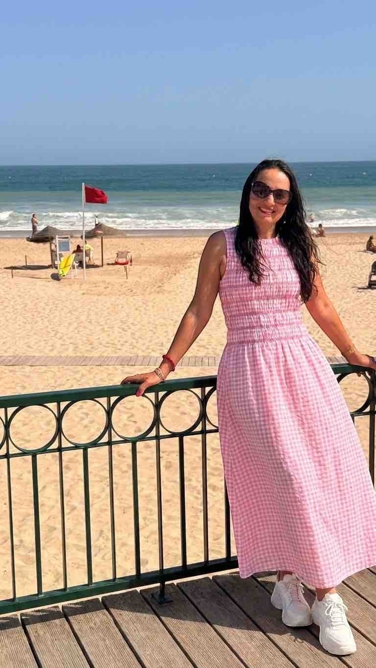 Antique Rose Boutique pink gingham maxi dress with a smocked bodice worn by a woman on a sunny beach boardwalk